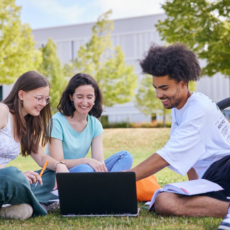 Drei Studierende sitzen auf dem Rasen, schauen auf einen Laptop und diskutieren. Ein Fahrrad steht im Hintergrund.