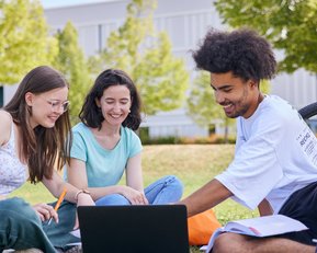 Drei Studierende sitzen auf dem Rasen, schauen auf einen Laptop und diskutieren. Ein Fahrrad steht im Hintergrund.