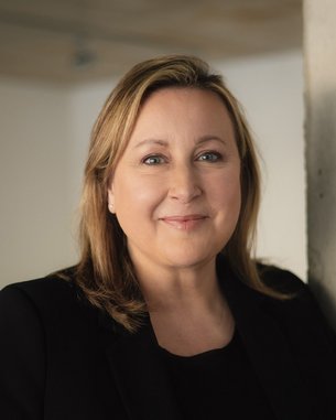 Eine Frau mit blonden Haaren und einem freundlichen Gesichtsausdruck steht vor einer hellen Wand. Sie trägt ein schwarzes Oberteil und hat eine natürliche Pose.