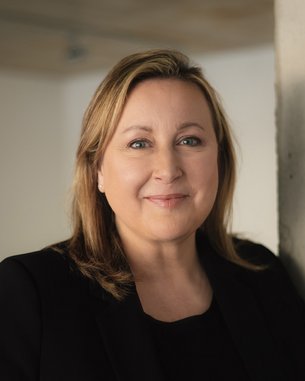 Eine Frau mit blonden Haaren und einem freundlichen Gesichtsausdruck steht vor einer hellen Wand. Sie trägt ein schwarzes Oberteil und hat eine natürliche Pose.