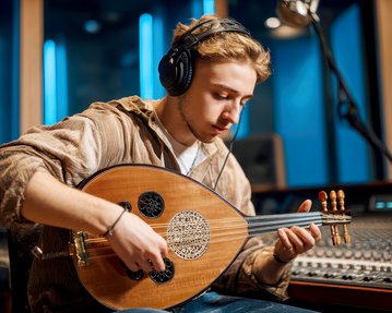 Ein junger Mann spielt eine Oud in einem Tonstudio, während er Kopfhörer trägt. Im Hintergrund sind Mischpult und Studiolichter sichtbar.