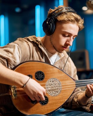 A young man is playing an oud in a recording studio while wearing headphones. In the background, a mixing console and studio lights are visible.