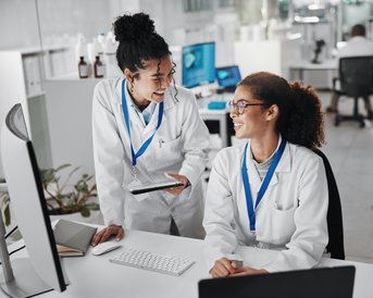 Zwei Wissenschaftlerinnen in Laborkitteln stehen an einem Schreibtisch, lächeln und diskutieren über ein Tablet.