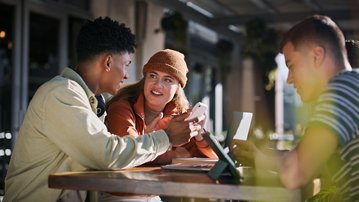 Vier junge Erwachsene sitzen an einem Tisch im Freien, unterhalten sich und nutzen ihre Smartphones. Ein Laptop und Notizen liegen auf dem Tisch.