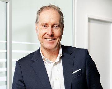 Prof. Schottler in einem dunklen Anzug steht lächelnd an einem Tisch, mit einer weißen Hemd und einer Brusttasche. Helle, moderne Büroumgebung.