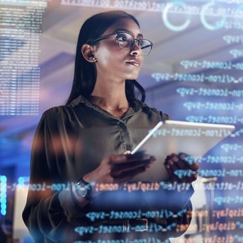 A woman with glasses is holding a tablet and looking at digital data and programming code on a screen in a modern office.