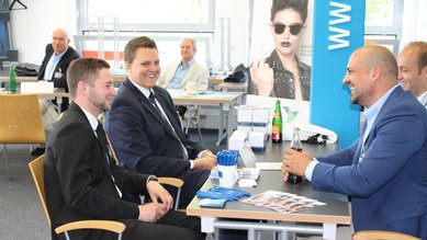 Vier Männer sitzen an einem Tisch und führen ein Gespräch. Im Hintergrund sind weitere Personen und Werbematerialien sichtbar.