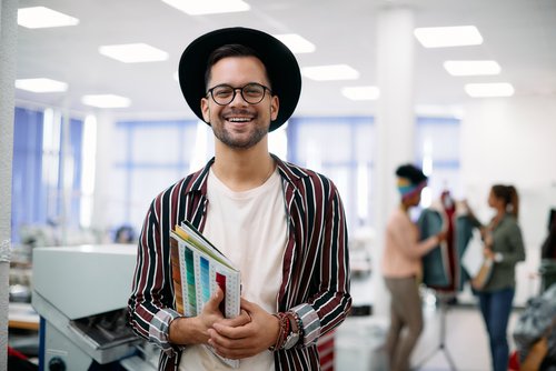 Young man with glasses and a hat, standing in a creative work environment and holding several color samples.
