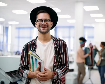 Junger Mann mit Brille und Hut, der in einem kreativen Arbeitsumfeld steht und mehrere Farbmuster in der Hand hält.