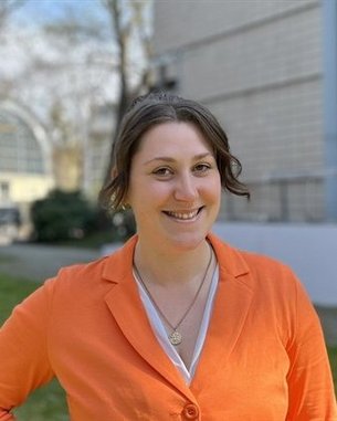 Eine Person mit braunen Haaren trägt ein orangefarbenes Jackett und lächelt. Im Hintergrund sind Bäume und ein Gebäude sichtbar.