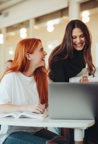 Eine Gruppe von Studierenden sitzt in einem modernen Klassenraum. Zwei Frauen lachen und unterhalten sich, während eine dritte Frau am Laptop arbeitet. Weitere Studierende sind im Hintergrund mit Laptops und Notizen beschäftigt.