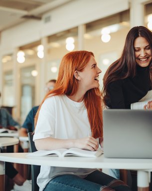 Eine Gruppe von Studierenden sitzt in einem modernen Klassenraum. Zwei Frauen lachen und unterhalten sich, während eine dritte Frau am Laptop arbeitet. Weitere Studierende sind im Hintergrund mit Laptops und Notizen beschäftigt.