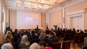Ein Konferenzraum mit vielen Teilnehmern, die auf eine Präsentation warten. Ein Bildschirm zeigt das Wort "DAV".