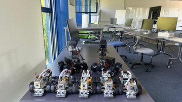 Sechs Roboterfahrzeuge stehen auf einem Tisch in einem modernen Raum mit Computerarbeitsplätzen im Hintergrund.