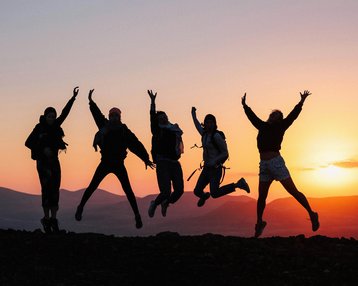 Sechs Personen springen vor einem Sonnenuntergang, silhouettiert gegen den Himmel, mit erhobenen Armen und fröhlichen Ausdrücken.
