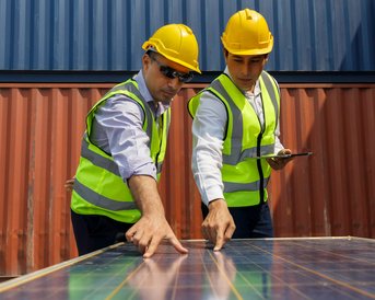 Two men in safety vests and helmets are examining a solar panel together and discussing its details.