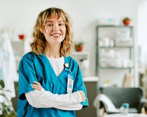 Eine lächelnde Krankenschwester in blauer Uniform steht mit verschränkten Armen in einem modernen Gesundheitsumfeld.
