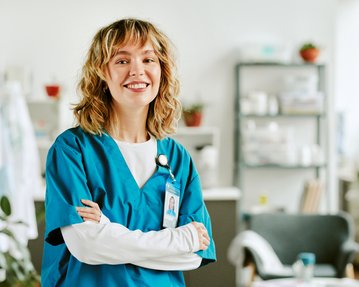 Eine lächelnde Krankenschwester in blauer Uniform steht mit verschränkten Armen in einem modernen Gesundheitsumfeld.