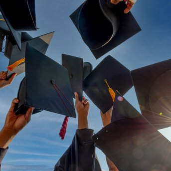 Hände halten Abschlussmützen in die Luft, während sie den Abschluss feiern. Der Himmel ist klar und sonnig.