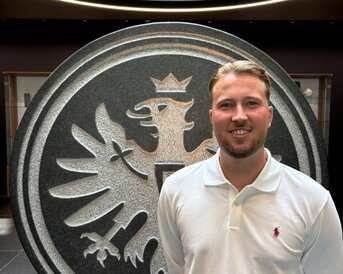 Ein Mann steht vor einem großen, runden Emblem mit einem Adler und einer Krone, das auf einem Steinsockel platziert ist.