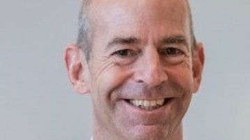 Ein Mann mit kurzen, grauen Haaren und einem freundlichen Lächeln trägt einen schwarzen Anzug und ein weißes Hemd. Der Hintergrund ist neutral und unscharf, was den Fokus auf das Gesicht des Mannes lenkt.