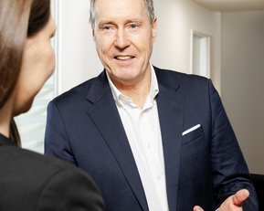 Ein Mann in einem Anzug spricht mit einer Frau, die ihm zugewandt ist. Beide stehen an einem Empfangstresen in einem modernen Raum.