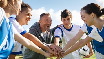 Eine Gruppe von jungen Fußballspielern und einem Trainer versammelt sich im Freien, um sich mit den Händen in der Mitte zu motivieren.