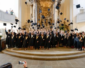Graduates in black gowns and caps stand in a church, tossing their caps into the air. They happily pose in front of an ornate wall adorned with sculptures. Some spectators can be seen in the background.