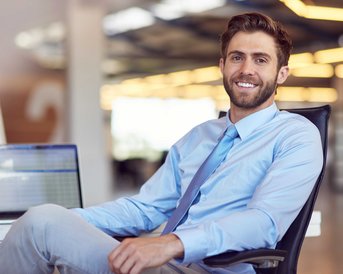 A smiling man in a light blue shirt and a tie is sitting at a desk. In the background, a laptop is visible, and the surroundings appear modern and bright.
