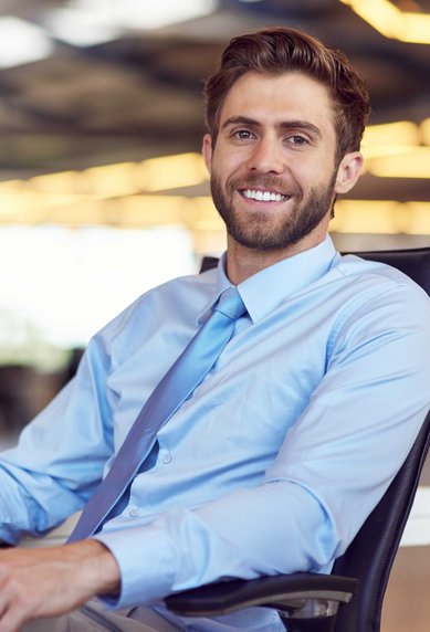 A smiling man in a light blue shirt and a tie is sitting at a desk. In the background, a laptop is visible, and the surroundings appear modern and bright.