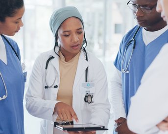 Eine Ärztin erklärt einem Team von vier medizinischen Fachkräften Informationen auf einem Tablet in einem modernen Krankenhausumfeld.