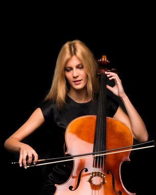 A woman with long blonde hair is playing a cello. She sits in a dimly lit room, holding the bow in her right hand while pressing the strings with her left.