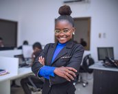 Eine lächelnde Frau in einem schwarzen Anzug steht mit verschränkten Armen in einem modernen Büro. Sie trägt eine blaue Bluse und Brille, während im Hintergrund mehrere Personen an Computern arbeiten.