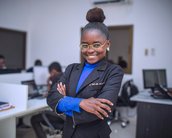Eine lächelnde Frau in einem schwarzen Anzug steht mit verschränkten Armen in einem modernen Büro. Sie trägt eine blaue Bluse und Brille, während im Hintergrund mehrere Personen an Computern arbeiten.