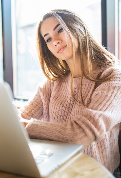 A young woman sits at a table, focused on her laptop, in a bright room with large windows.