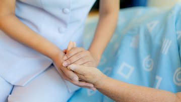 Eine Pflegekraft hält die Hand eines Patienten, der auf einem Bett mit blauer Bettwäsche sitzt.