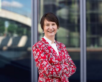 Frau mit kurzem Haar trägt einen roten Blazer mit Blumenmuster und steht lächelnd vor einer modernen Glasfassade.