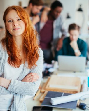 Eine lächelnde Frau mit roten Haaren steht im Vordergrund, während mehrere Personen im Hintergrund an einem Tisch arbeiten.