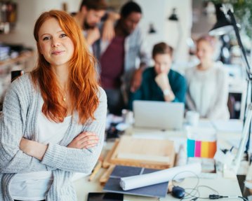 Eine lächelnde Frau mit roten Haaren steht im Vordergrund, während mehrere Personen im Hintergrund an einem Tisch arbeiten.