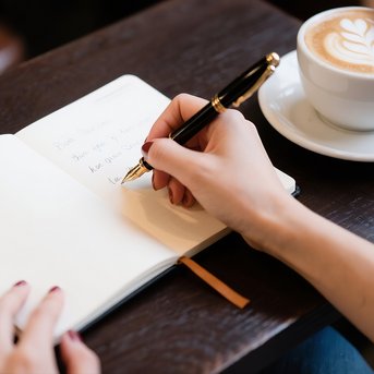 Eine Hand schreibt in ein offenes Notizbuch, während eine Tasse Kaffee auf einem Tisch steht.