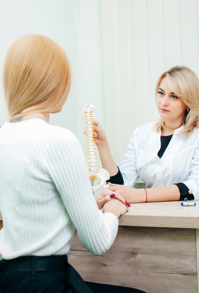 Eine Ärztin erklärt einer Patientin eine Wirbelsäule, während sie an einem Schreibtisch sitzt. Im Hintergrund sind Röntgenbilder zu sehen.