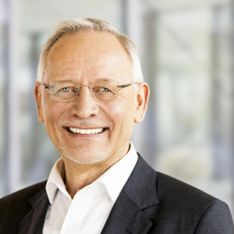 Ein älterer Mann mit Brille und grauen Haaren lächelt freundlich in einem formellen Anzug vor einem unscharfen Hintergrund.