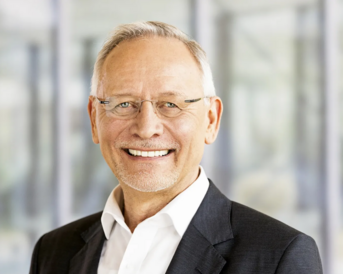 Ein älterer Mann mit Brille und grauen Haaren lächelt freundlich in einem formellen Anzug vor einem unscharfen Hintergrund.