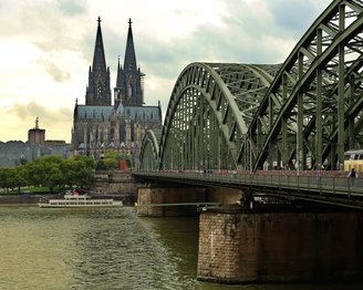 Die Hohenzollernbrücke über den Rhein mit Blick auf den Kölner Dom im Hintergrund. Die Brücke hat eine grüne Stahlkonstruktion und ist von Wasser und Bäumen umgeben.