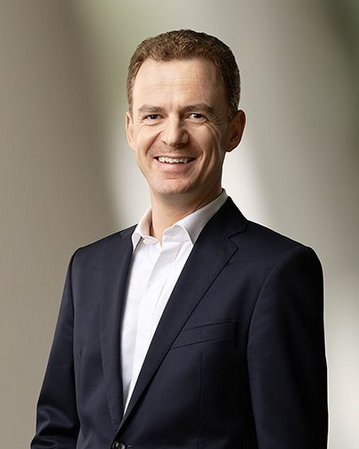 Ein Mann in einem dunklen Anzug mit weißem Hemd lächelt freundlich. Der Hintergrund ist unscharf und hell.