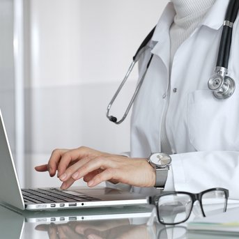 Eine Person in einem weißen Kittel tippt auf einem Laptop, während ein Stethoskop um den Hals hängt. Eine Brille liegt auf dem Tisch.