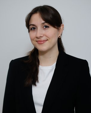 Junge Frau mit langen, braunen Haaren, die in einem schwarzen Blazer und einem weißen Oberteil lächelt. Hintergrund ist neutral.