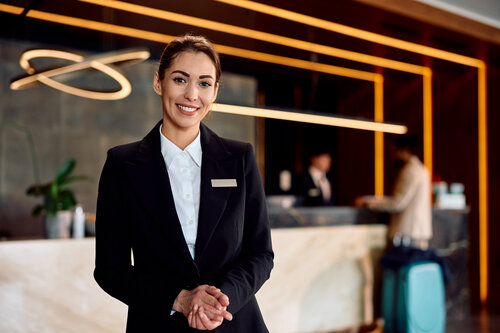 Eine lächelnde Hotelangestellte in formeller Kleidung steht am Empfangstresen, während Gäste im Hintergrund einchecken.