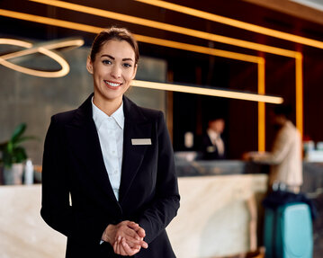 Eine lächelnde Hotelangestellte in formeller Kleidung steht am Empfangstresen, während Gäste im Hintergrund einchecken.