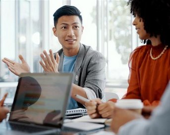 A man is explaining something in a meeting while several people are sitting around a table with laptops and notes.